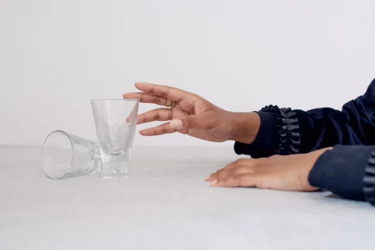 Eine Hand greift nach einem kleinen, durchsichtigen Glas, das auf einem Tisch steht. Daneben liegt ein weiteres Glas seitlich auf der Oberfläche. Die Szene ist schlicht gehalten, mit hellem Hintergrund und Fokus auf die Hände und die Gläser.