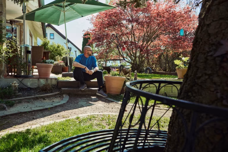 Mann sitzt entspannt im sonnigen Garten vor einem Wohnhaus, umgeben von Pflanzen, Bäumen und ruhiger Atmosphäre.