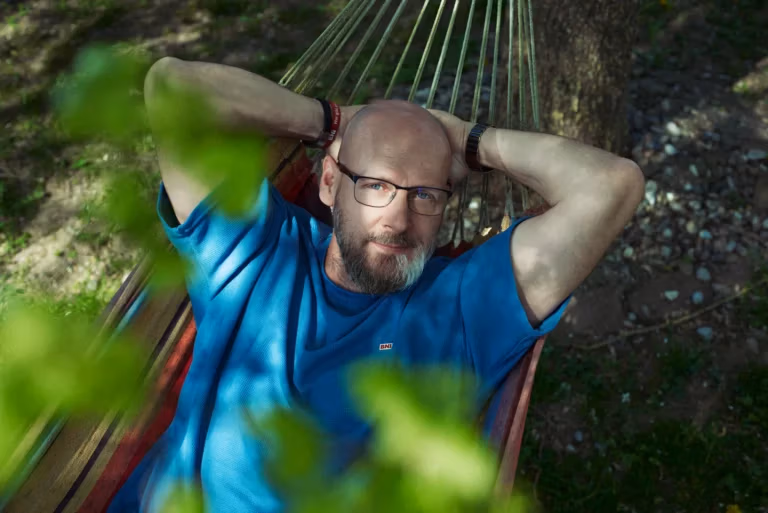 Mann mit Brille entspannt in einer Hängematte im Grünen, aufgenommen im natürlichen Sonnenlicht zwischen unscharfen Blättern.