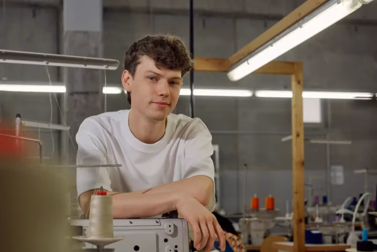 Porträt eines jungen Mannes in den Produktionshallen eines nachhaltigen Textilunternehmens, umgeben von Nähmaschinen, Stoffen und Arbeitsplätzen. Er blick leicht Frech in die Kamera und macht einen selbstbewussten Eindruck.