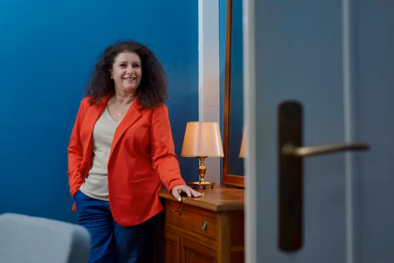 Porträt einer Frau im roten Blazer angelehnt an eine Kommode aus Holz in einem stilvoll eingerichteten Innenraum mit farbenfrohen Sesseln, großen Fenstern und entspannter Atmosphäre. Ihr Blick ist gelöst, energetisch und zur Kamera gerichtet.