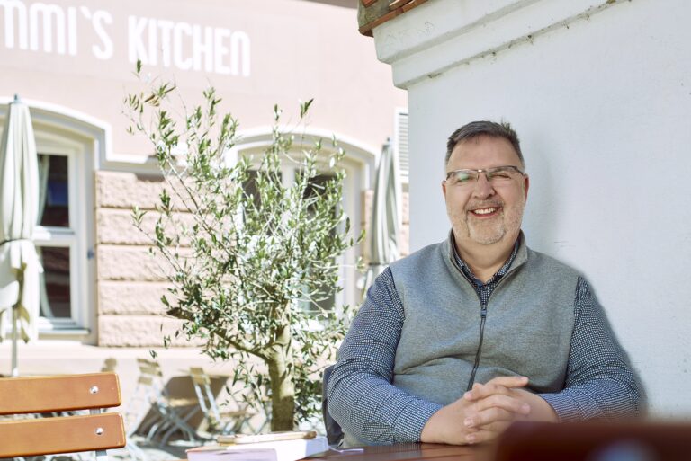 Andreas Becker sitzt lächelnd an einem Tisch in der Augsburger Altstadt. Der Unternehmensberater trägt ein graues Fleece-Gilet über einem gemusterten Hemd und lehnt entspannt an einer hellen Mauer. Im Hintergrund sind ein Olivenbaum, Café-Mobiliar und die Fassade eines Restaurants zu sehen, wodurch eine freundliche und entspannte Atmosphäre entsteht.